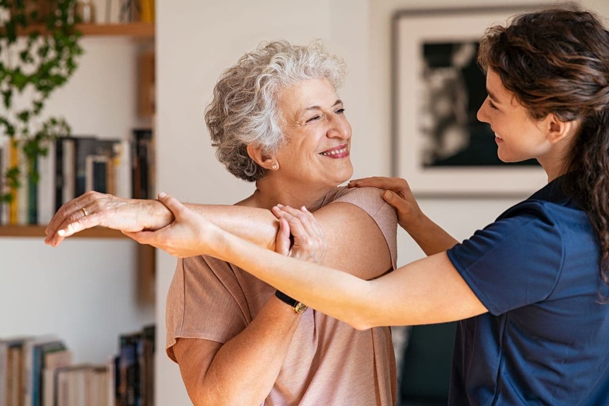 A female therapist assisting an older woman with a shoulder stretch.