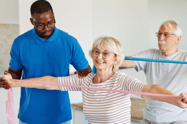 A man giving instructions to an older woman performing an exercise with a resistance band.