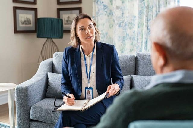 A female support worker conversing with an older man while taking notes