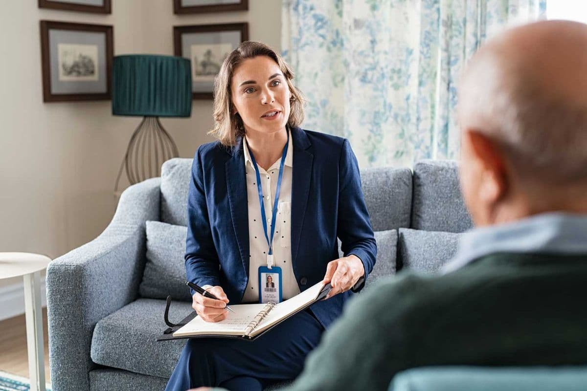 A female support worker conversing with an older man while taking notes