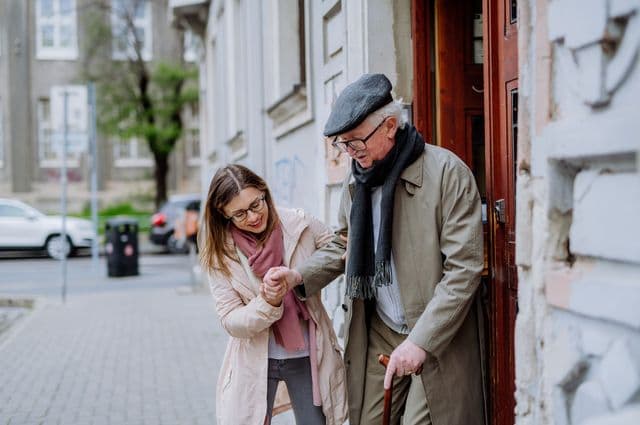 A young woman helping an older man out the door for a walk on the street.