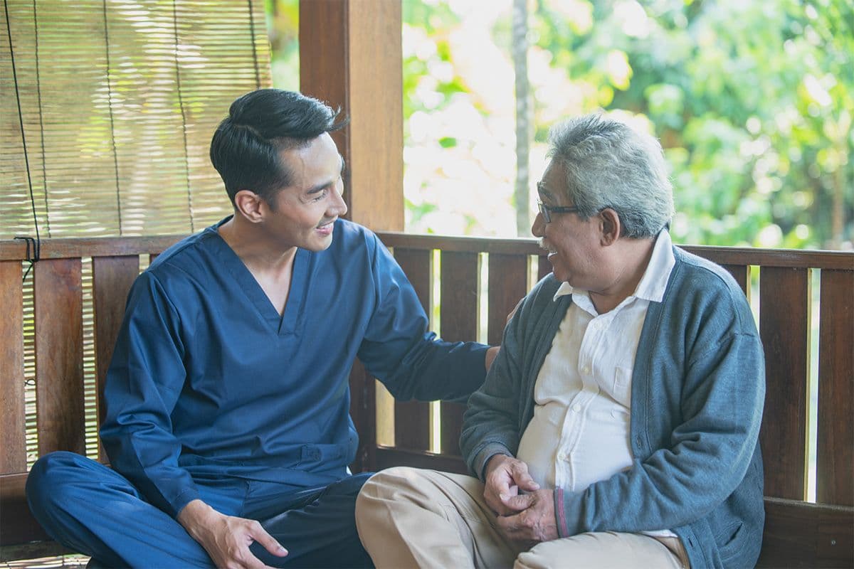 A healthcare worker in blue scrubs sitting next to an older man on a wooden bench