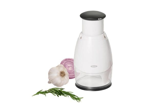 A manual food chopper alongside an onion, garlic, and rosemary