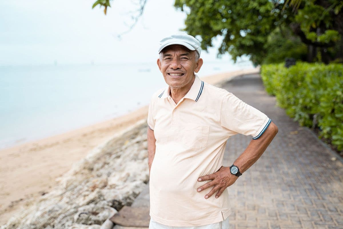 An older man standing on a paved pathway near a beach, wearing a light-coloured polo shirt and pants