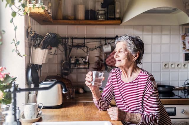 An older woman with short grey hair leans against a wooden kitchen counter holding a glass of water.