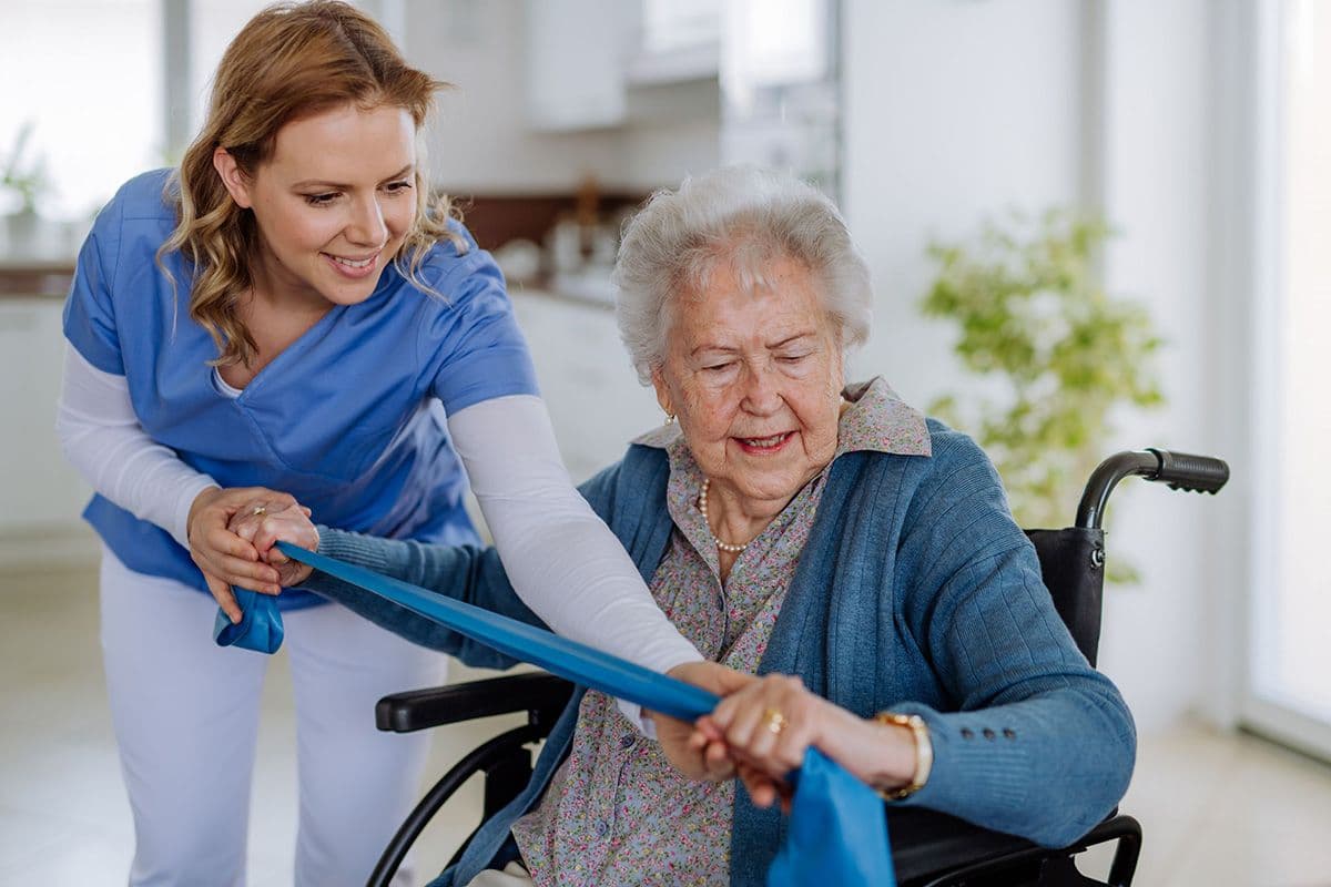 A healthcare professional in blue scrubs assists an older woman in a wheelchair with a resistance band exercise