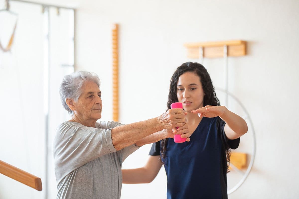 An older woman holding a pink dumbbell is assisted by a physiotherapist during a physical therapy session