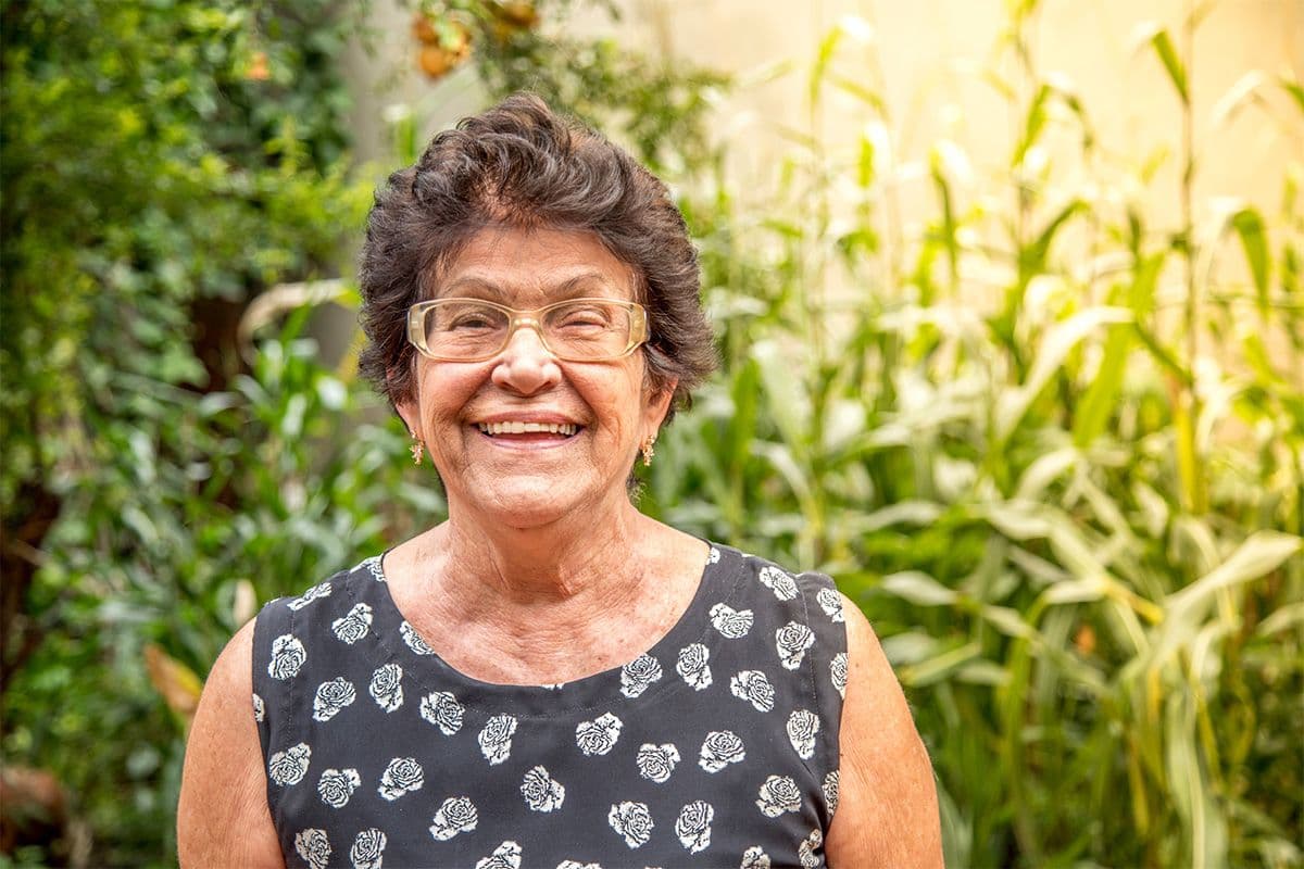 An older woman standing outdoors in a garden surrounded by lush green plants.