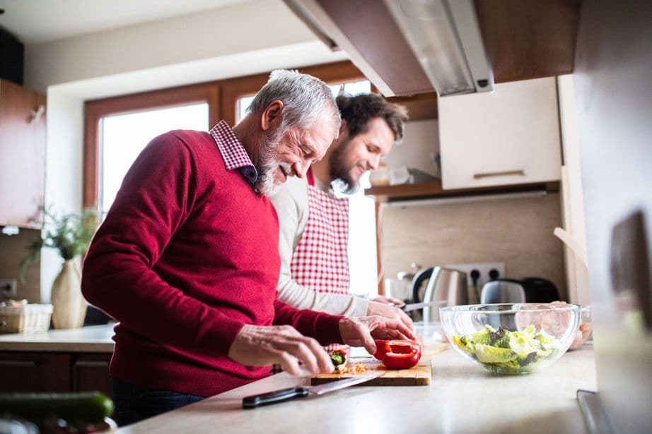 An older man happily preparing food in the kitchen with a younger man