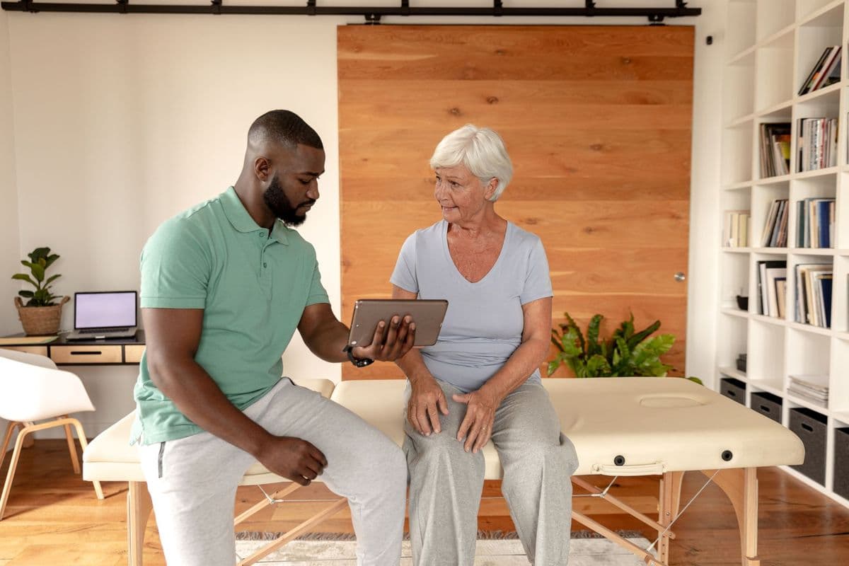 A physiotherapist explaining something to an older woman patient, showing her something on his tablet.
