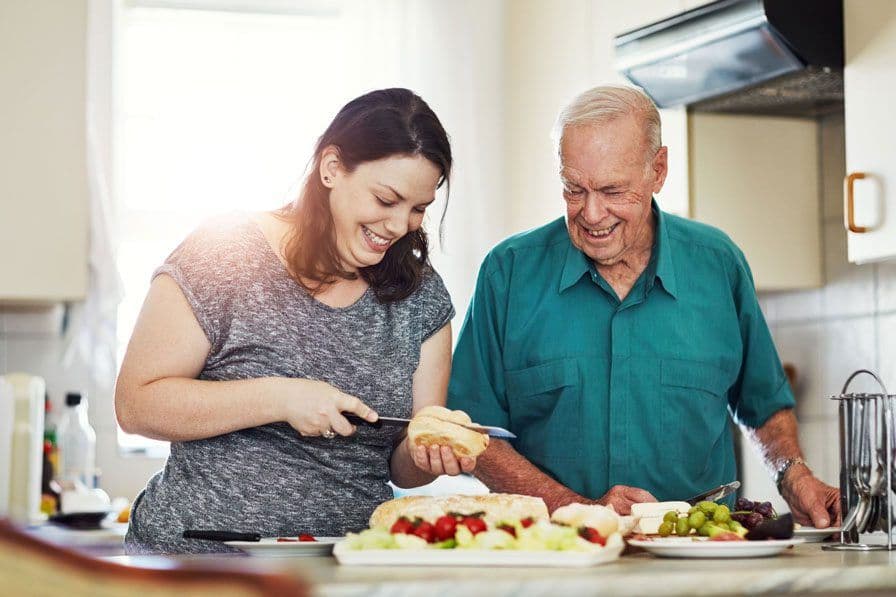 An older man happily preparing food in the kitchen with his adult daughter
