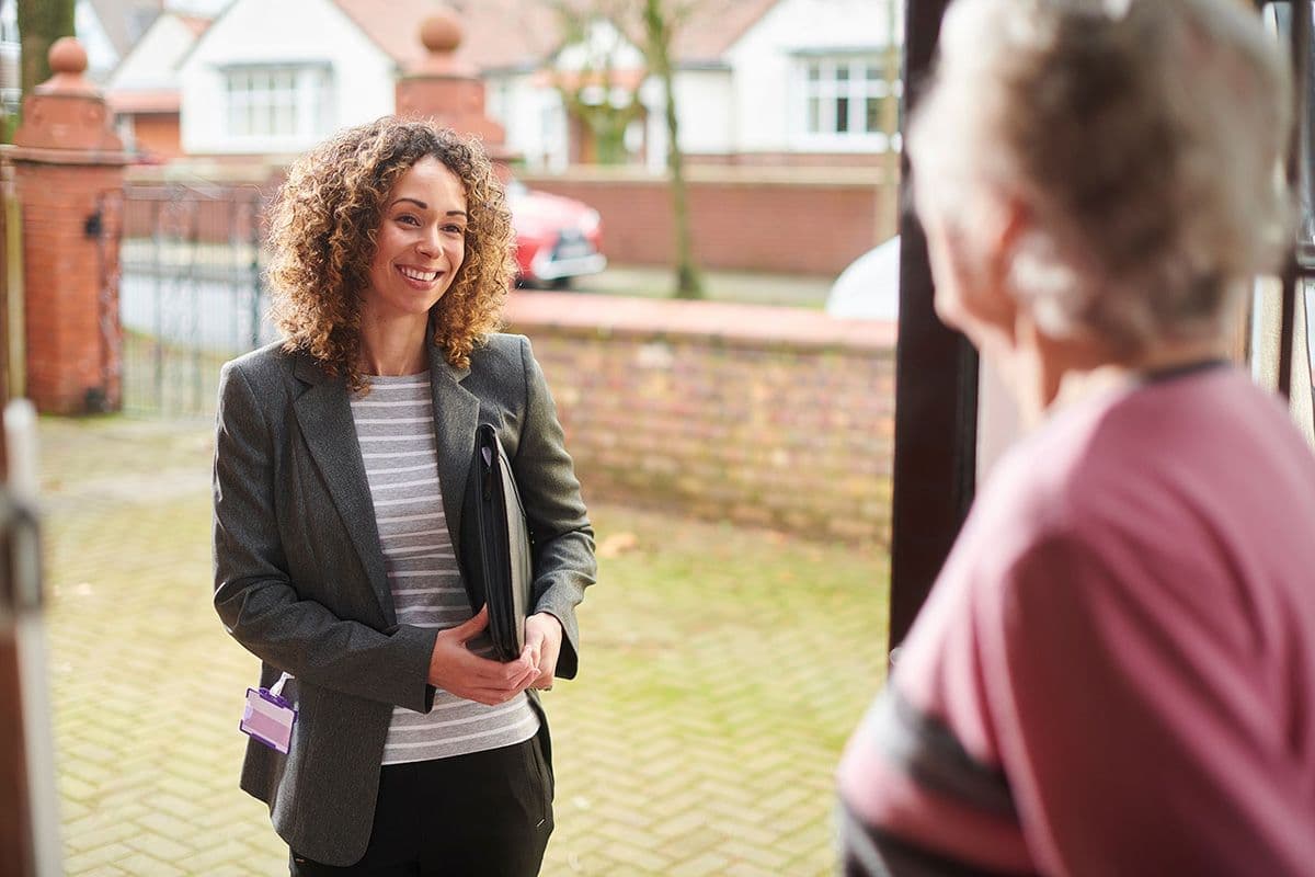 Two people interacting at the entrance of a house. One is holding a folder and standing outside, the other inside the doorway