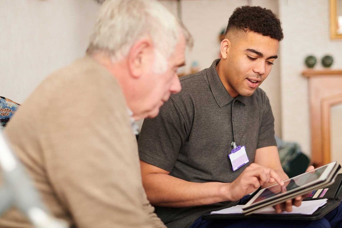 A male support worker conversing with an older man while recording notes on an iPad