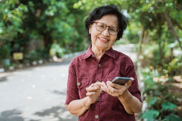 An older woman happily typing on her mobile phone while standing outside