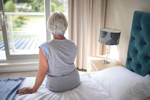 Older lady sat on the edge of a bed looking out of a window