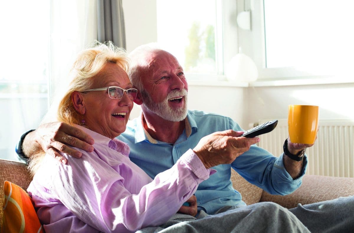 Happy senior couple watching television at home, sitting on sofa covered with blanket
