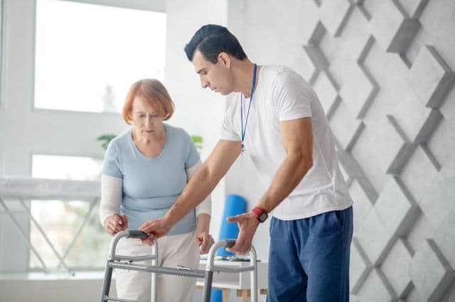 A male therapist assisting an older woman with a walking frame.