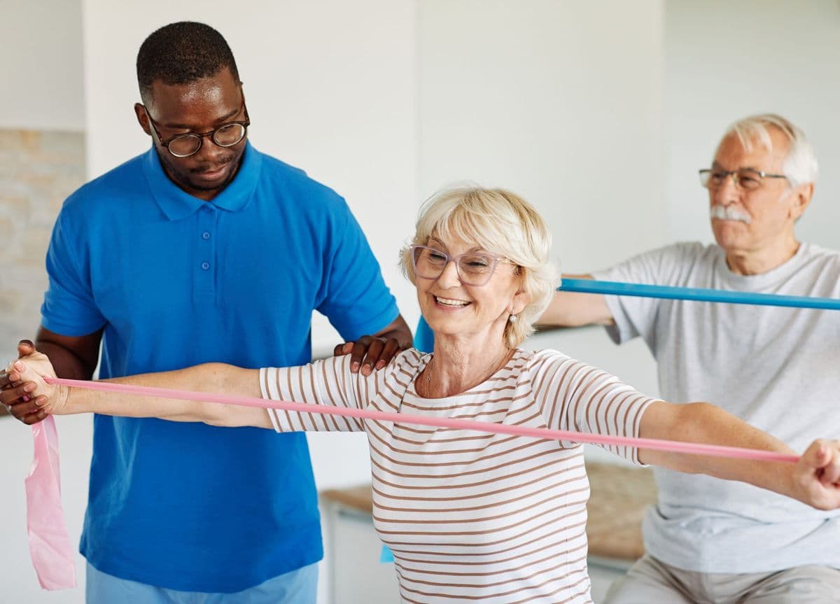 A man giving instructions to an older woman performing an exercise with a resistance band.
