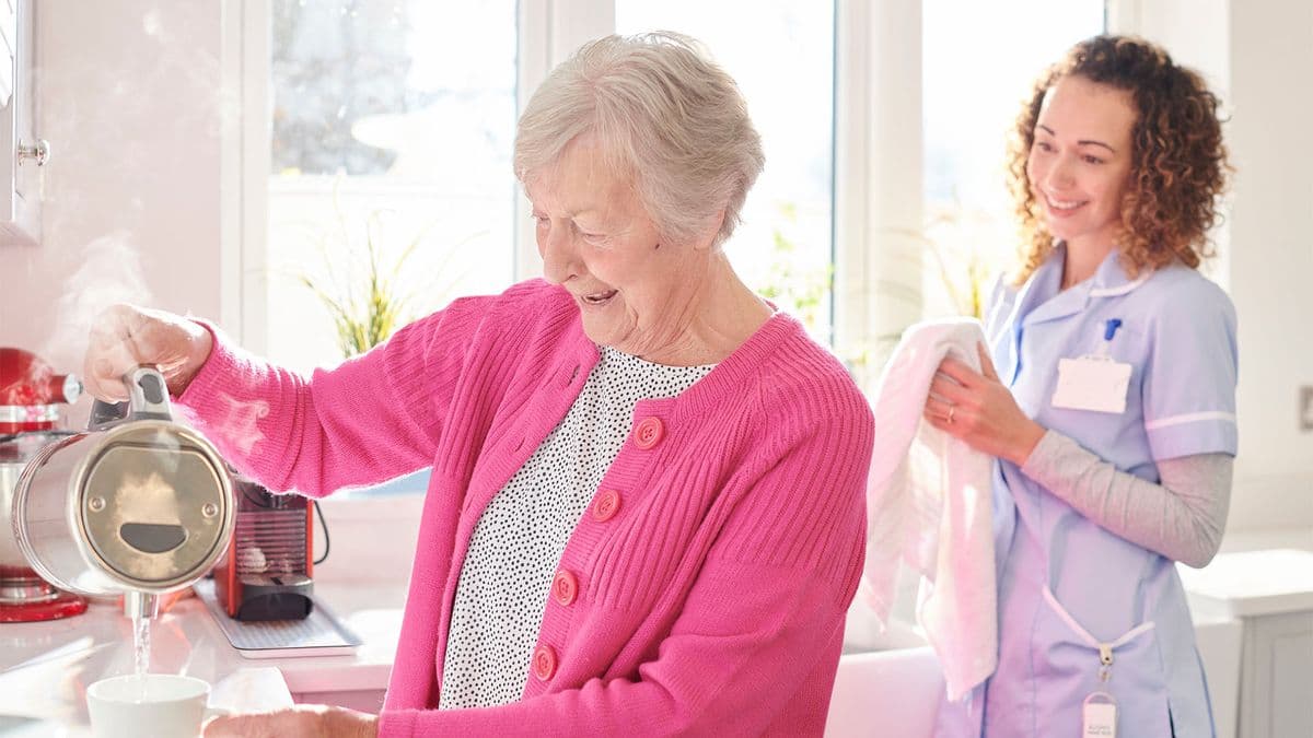 Older woman pouring water from a kettle while a support worker watches on