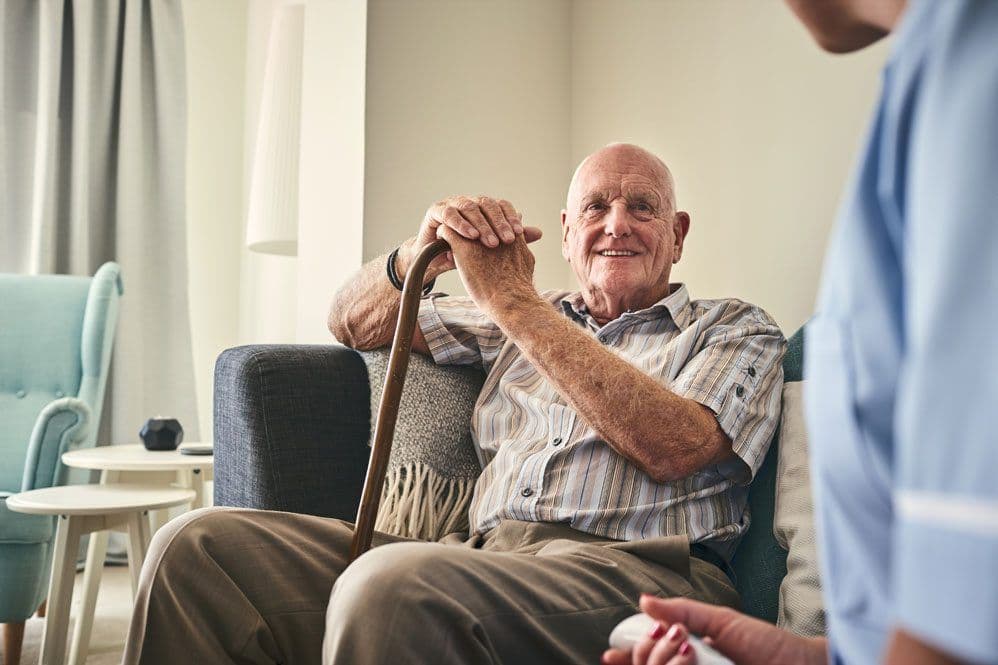 An older man sitting on a lounge and smiling at a support worker
