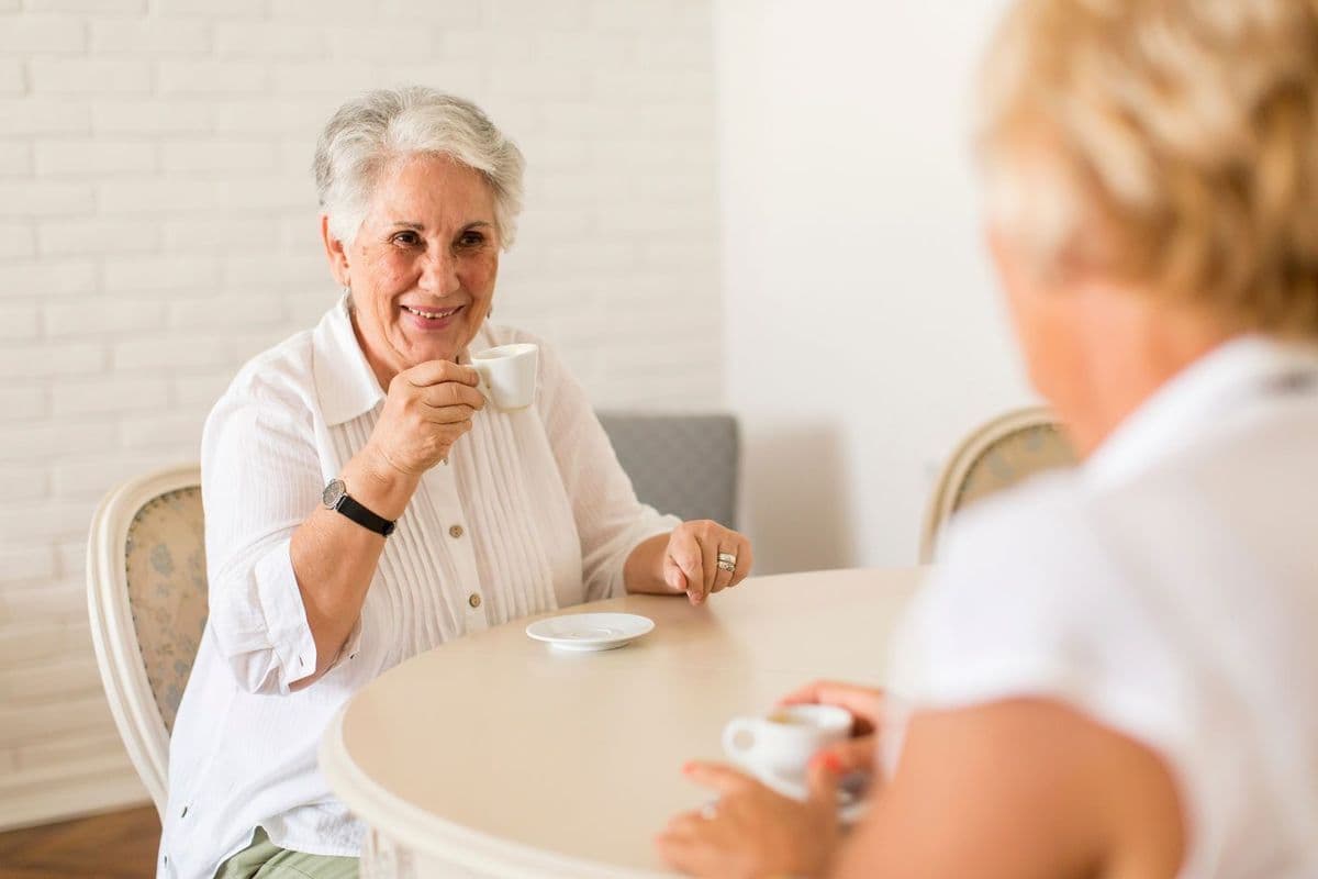 An older woman seated at a dining table and holding an espresso cup while smiling
