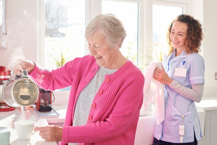 Older woman pouring water from a kettle while a support worker watches on