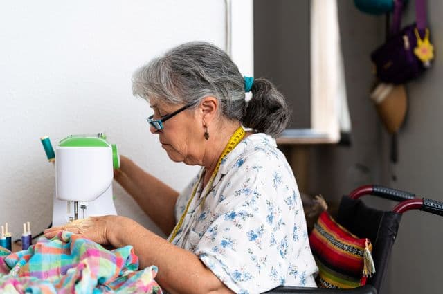 An older woman in wheelchair using a sewing machine.