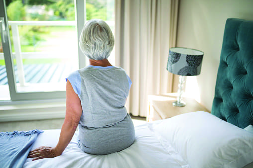 Older lady sat on the edge of a bed looking out of a window